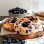 A close-up photo of three blueberry bagels on a brown surface, with a bowl of fresh blueberries in the background.