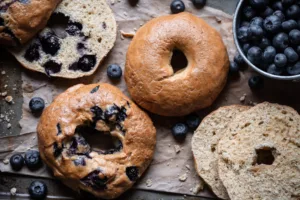 A close-up photo of three blueberry bagels on a brown surface, with a bowl of fresh blueberries in the background.