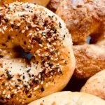 Close-up of freshly baked bagels, with one in the foreground topped with sesame seeds, black sesame seeds, poppy seeds, and dried onion flakes. The bagels appear golden brown and perfectly textured, showcasing a mix of soft and crispy surfaces.