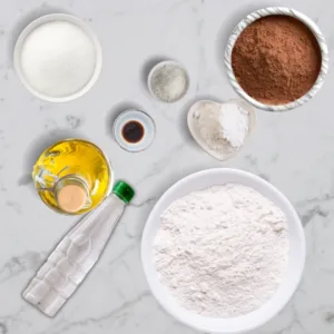 A flat lay of ingredients for wacky cake cupcakes, including flour, cocoa powder, sugar, baking soda, salt, vanilla extract, vinegar, and vegetable oil on a white marble surface.