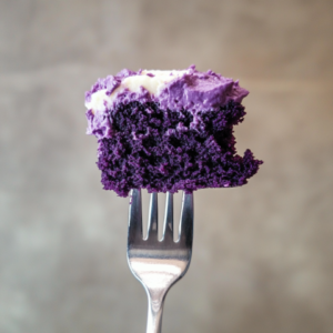 Close-up of a slice of purple velvet cake with smooth cream cheese frosting, showcasing its rich, vibrant purple layers and a decorative swirl of frosting on top.