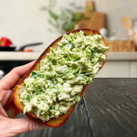 A hand holding a toasted slice of bread topped with creamy Green Goddess Tuna Salad, made with flaked tuna, avocado, herbs, and greens, in a modern kitchen setting.