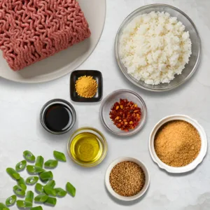 Flat-lay of key ingredients for a Korean-inspired BBQ beef bowl, including raw ground beef, cooked white rice, soy sauce, sesame oil, brown sugar, chili flakes, green onions, ginger powder, and sesame seeds, arranged on a light countertop