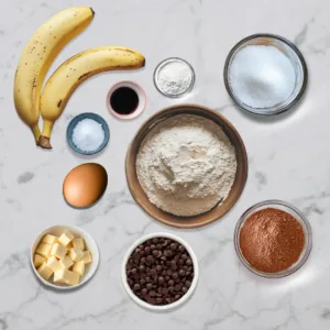 Ingredients for banana brownies, including ripe bananas, flour, cocoa powder, sugar, butter, egg, vanilla extract, salt, and chocolate chips.