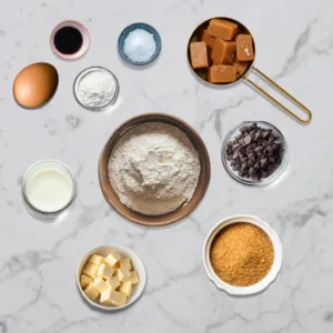 Ingredients for Knock You Naked Bars, including flour, caramel candies, chocolate chips, butter, brown sugar, egg, milk, vanilla extract, and salt on a marble countertop.